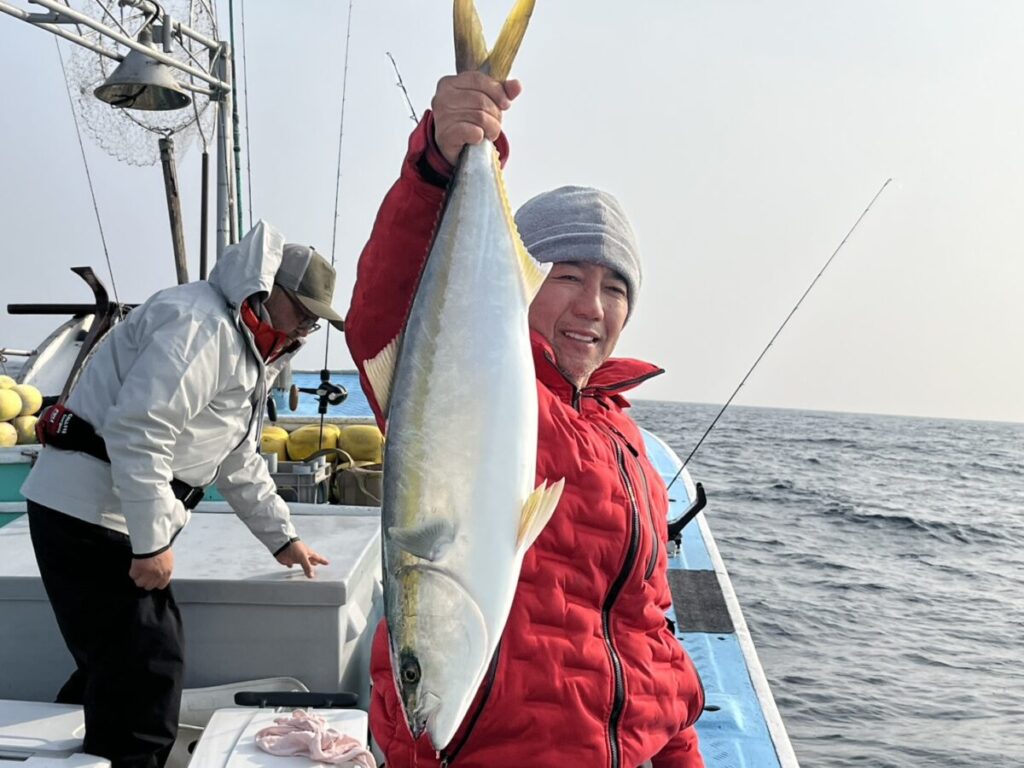 赤いジャケットにグレーのビーニーをかぶった人物が、ボートの上で笑顔で大きな魚を掲げている。船は曇り空の下、海の上にある。.