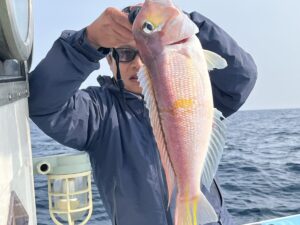 青いジャケットに眼鏡をかけた人物が、海と空を背景に、ボートに乗ったピンクと黄色の大きな魚を掲げている。.
