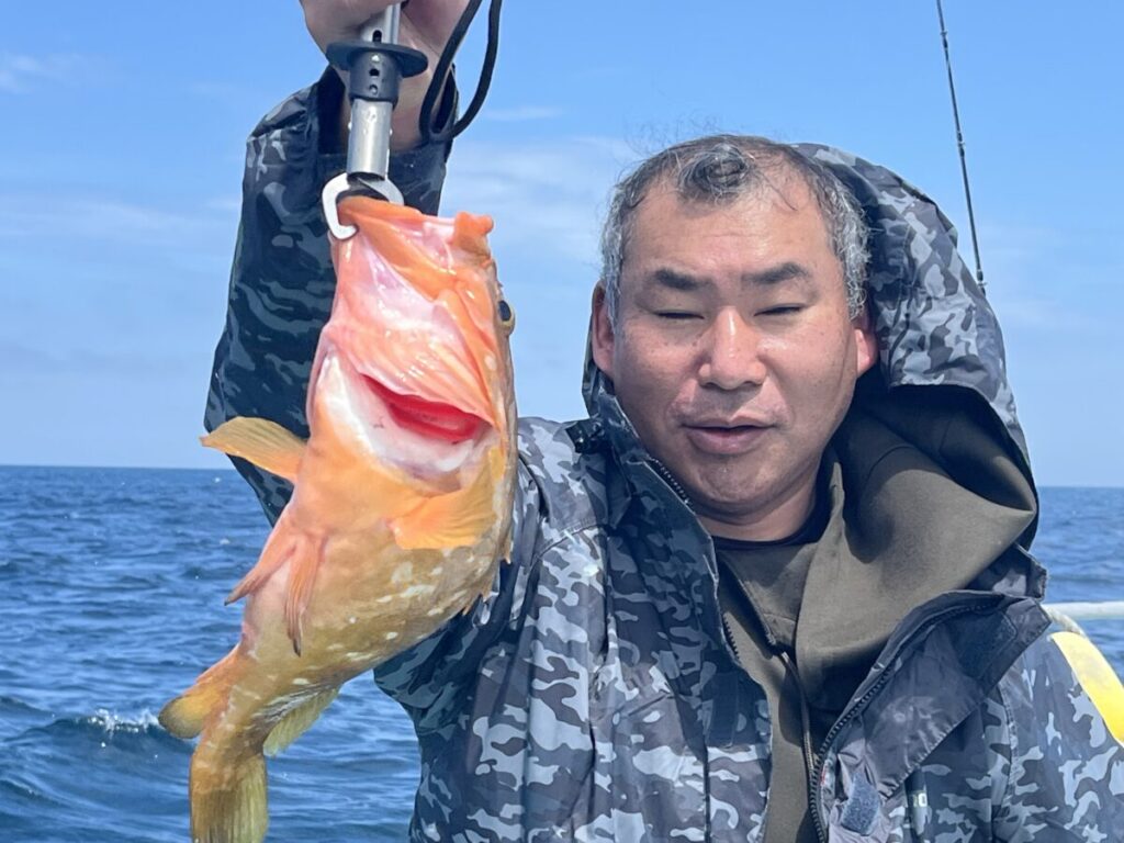 迷彩柄のジャケットを着た人物が、青空の下、海に浮かぶボートの上で、釣り針にかかった口を開けたオレンジ色の大きな魚を掲げている。.