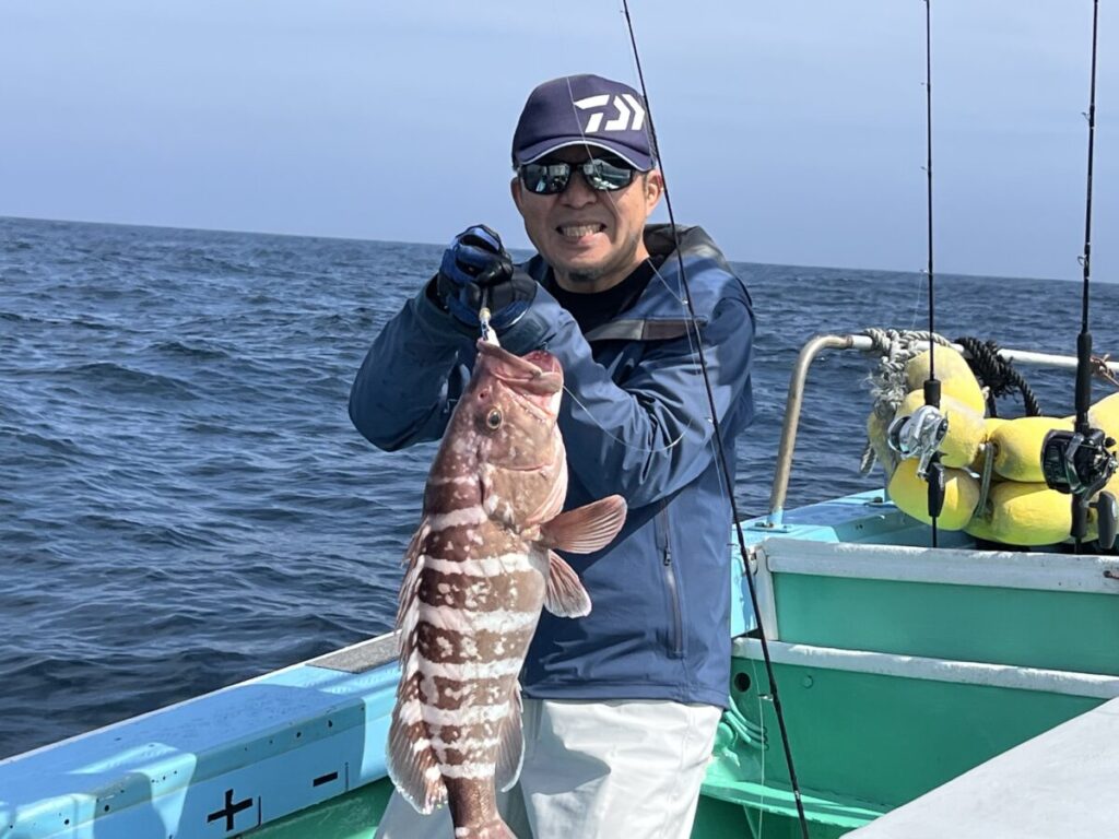 釣り上げた大きな縞模様の魚を掲げながら微笑むボートの男。サングラスをかけ、野球帽をかぶり、手袋をはめ、青いジャケットを着ている。背景には海と漁具が見える。.