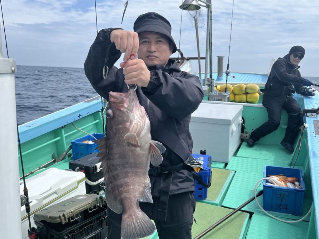 黒い服を着た人物がボートの上に立ち、両手で大きな魚を掲げている。ボートは水色で、漁具と黄色いブイで満たされている。海と空が見える。.