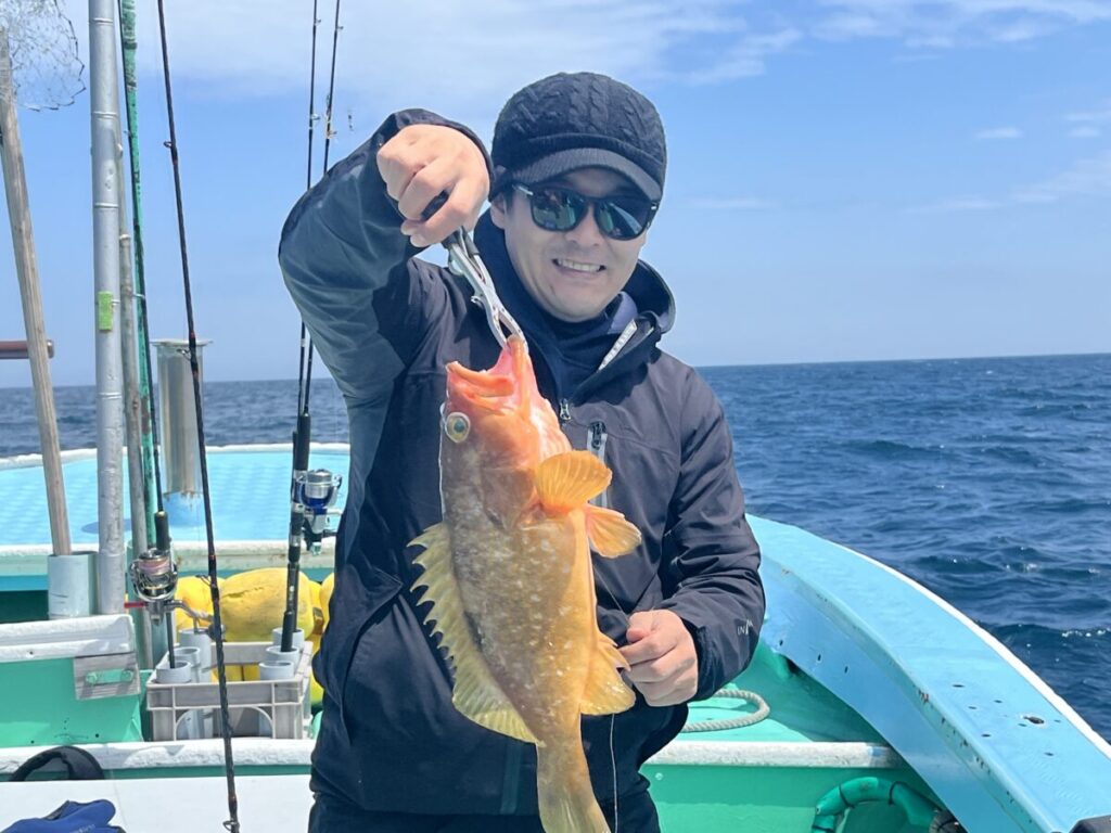 サングラスをかけ、黒いジャケットを羽織った人物が、オレンジ色の大きな魚を掲げながら微笑んでいる。.