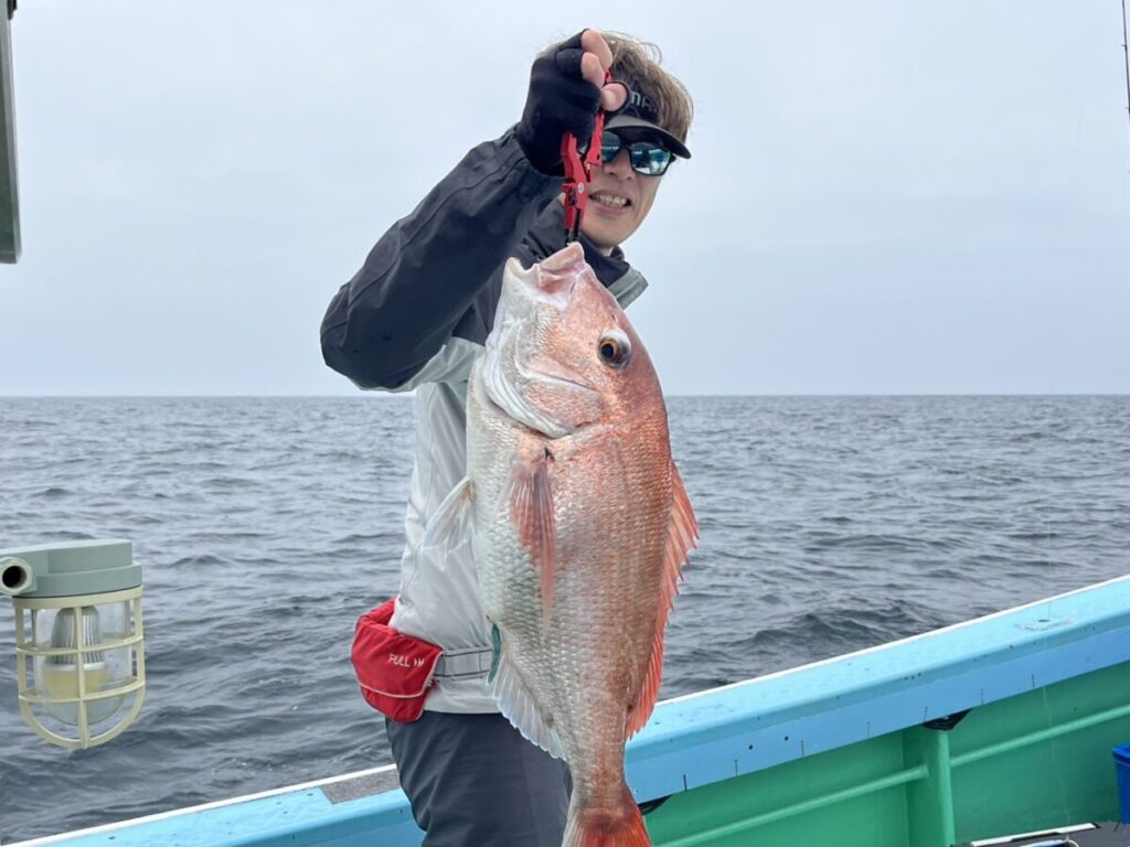 サングラスをかけ、手袋をはめた人物が、海上のボートの上で大きな魚を掲げ、カメラに向かって微笑んでいる。空は曇り、海は穏やかに見える。.