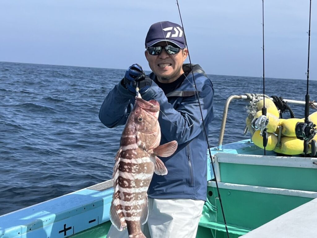 晴れ渡った空の下、釣り竿とブイを背景に、サングラスと帽子をかぶった人物が、海上の船上で大きな縞模様の魚を掲げて微笑んでいる。.