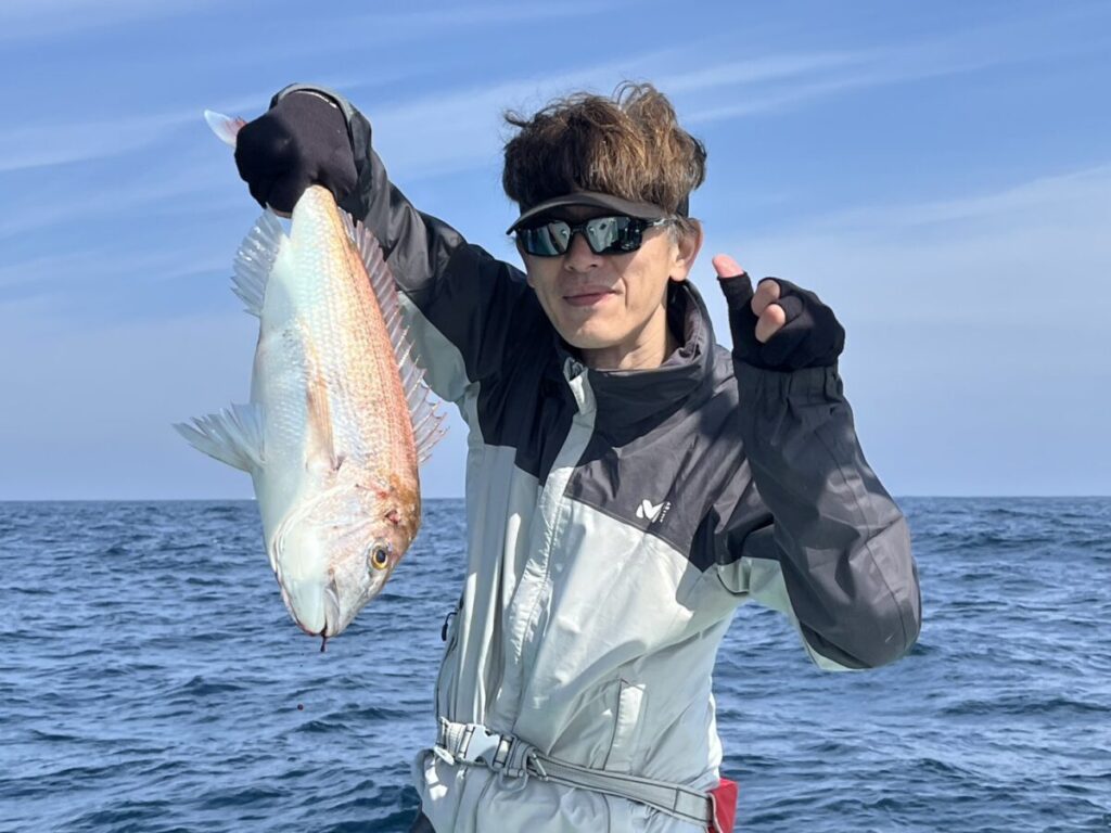 海と青空を背景に、サングラスをかけ、手袋をはめた人がボートの上で大きな魚を片手で持ち上げ、微笑んでいる。.