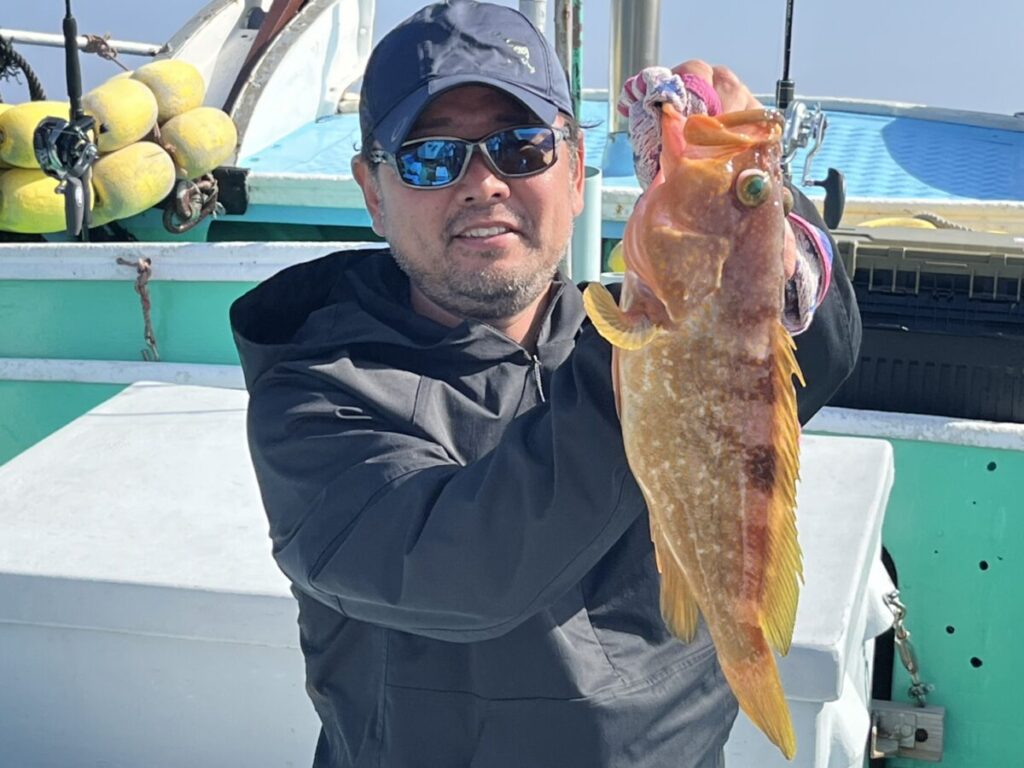 サングラスをかけ、黒っぽいジャケットを着た男が、オレンジがかった黄色の大きな魚をボートの上に掲げている。.