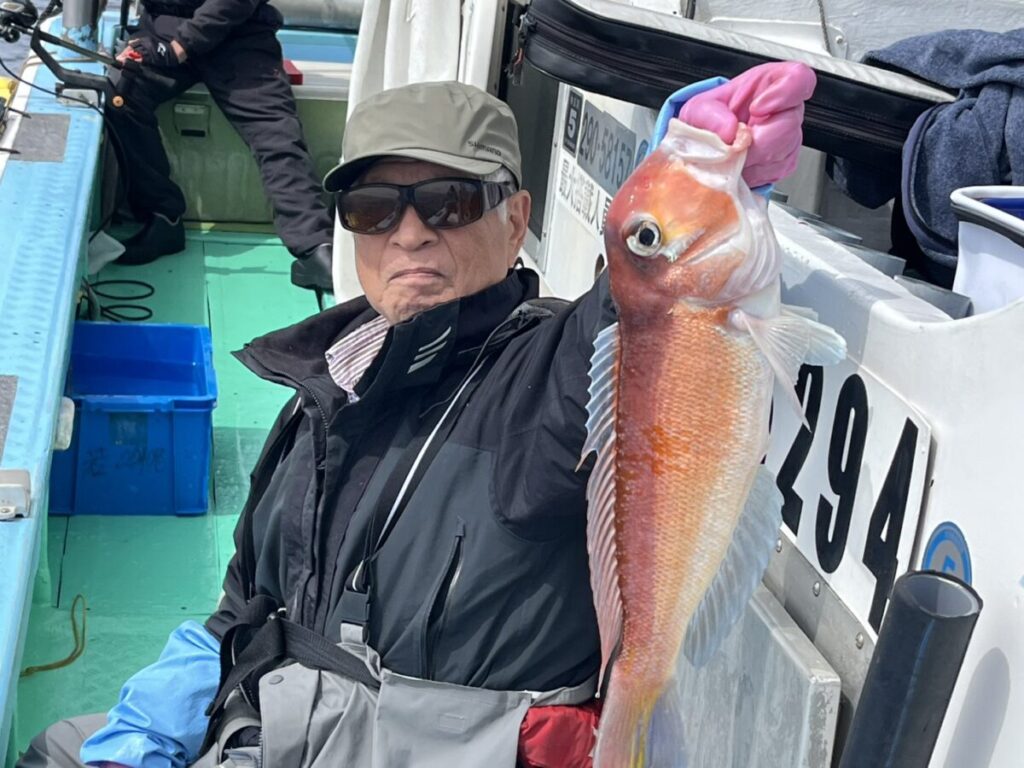 サングラスをかけ、帽子をかぶった年配の男性が、ボートの上で大きな魚を誇らしげに掲げてカメラに見せている。彼は釣りの道具を身につけ、手袋をしている。.