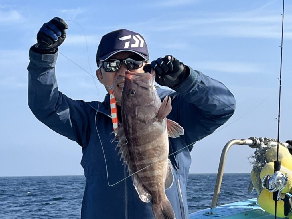 海と空を背景に、青い上着に帽子をかぶった人物が船の上で釣りたての魚を掲げている。.