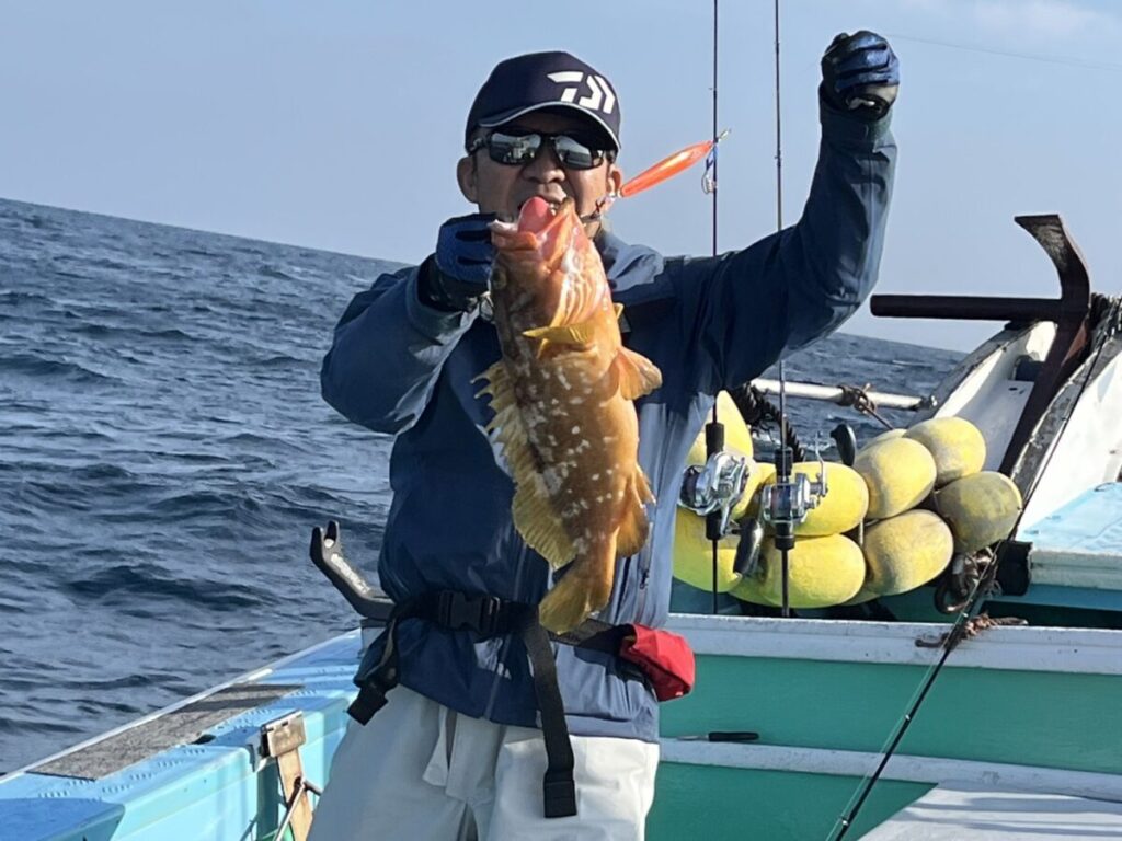 サングラスをかけ、帽子をかぶった人が、漁船の上で茶色がかった黄色の大きな魚を誇らしげに掲げている。.