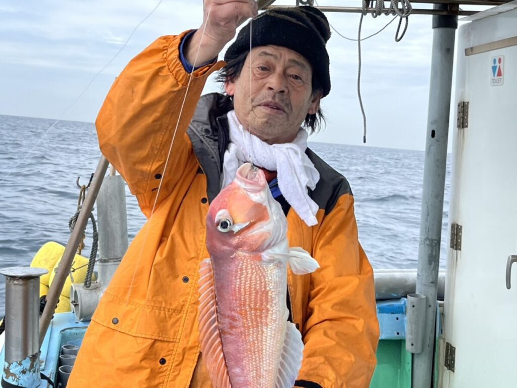 オレンジ色のジャケットに黒いビーニーをかぶった人物が、海上のボートの上で釣り糸を垂れるピンク色の大きな魚を掲げている。.