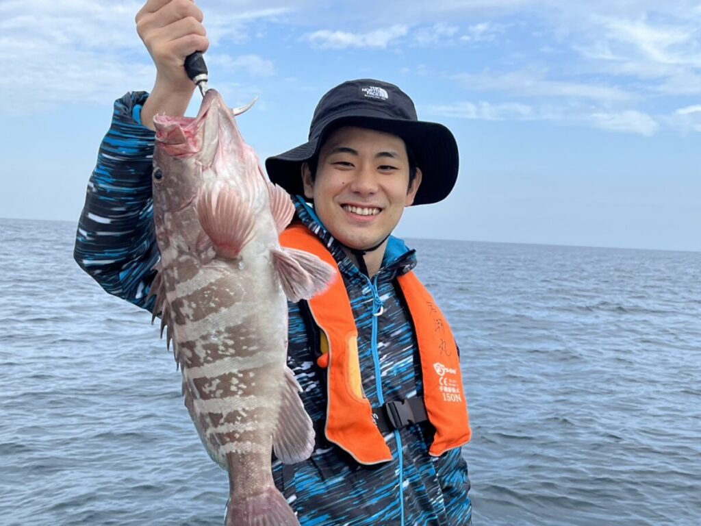 黒い帽子に青い上着、オレンジの救命胴衣を着た笑顔の人物が、大海原と空を背景に、ボートの上で釣りたての大きな魚を掲げている。.