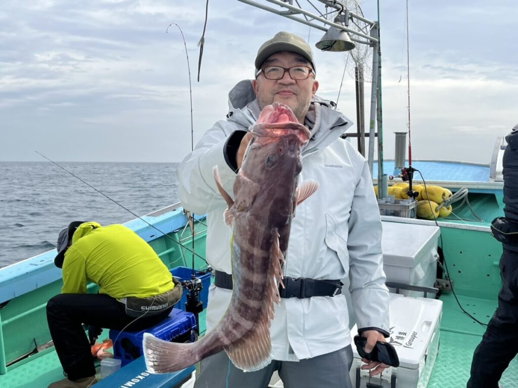 眼鏡をかけ、白いジャケットを着た人物がボートの上で大きな魚を誇らしげに抱えている。近くには釣り道具を持った黄色いジャケットの人物が座っている。背景には海と曇り空が見える。.