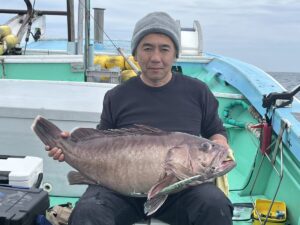 グレーのビーニーをかぶり、黒いシャツを着た男性が、釣りたての大きな魚を両手で抱えてボートに座っている。ボートには青と緑のディテールがあり、背景には漁具が見える。.