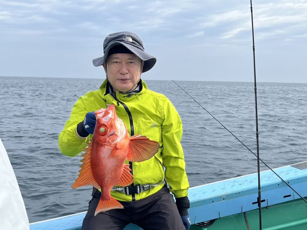 鮮やかな黄色のジャケットに帽子をかぶった人物がボートに乗り、海と曇り空を背景に大きな赤い魚を掲げている。.