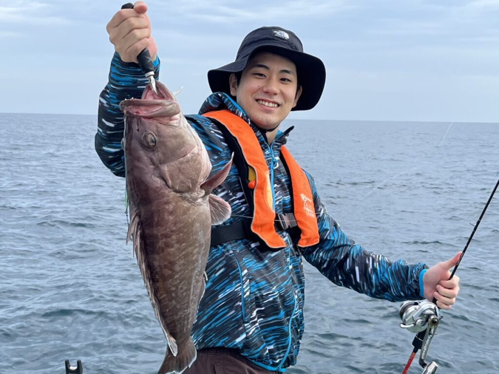 青い柄のジャケットとオレンジの救命胴衣を着た笑顔の人物が、穏やかな海と曇り空を背景に、海上でボートの上に立ちながら大きな魚を掲げている。.