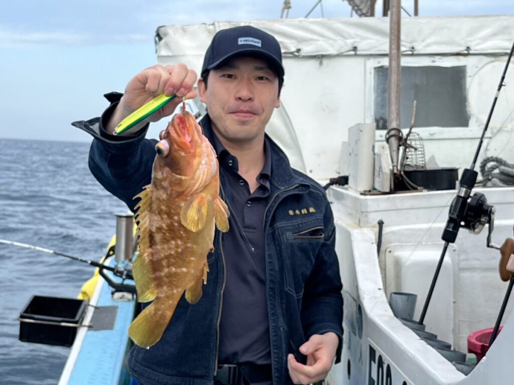 ボートの上に立つ男が、黄色い斑点のある茶色の大きな魚を掲げ、カメラに向かって微笑んでいる。背景には海と漁具が見える。.