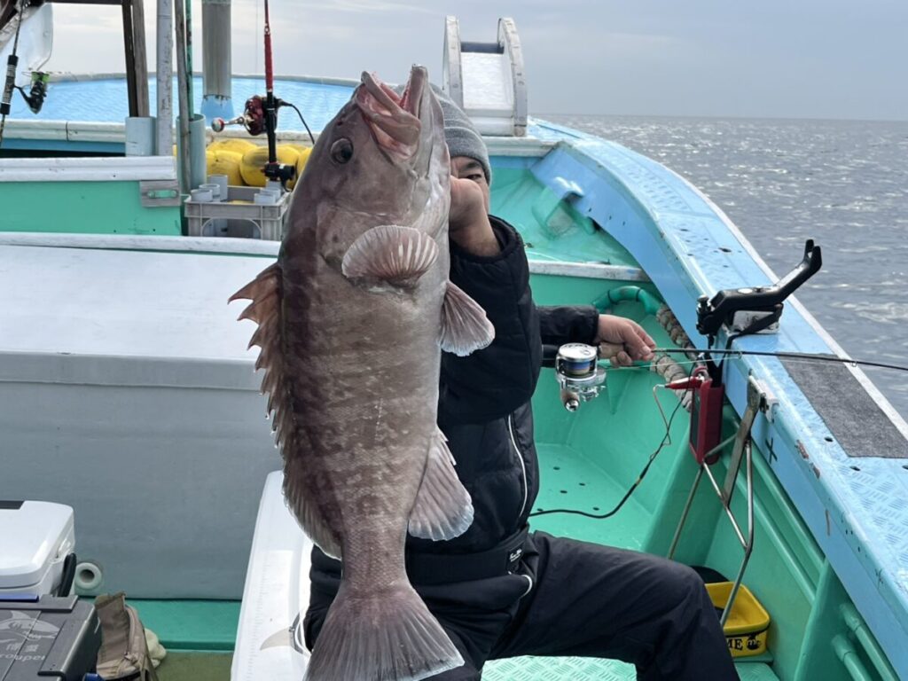 ターコイズブルーの漁船に乗った人が、顔を隠して釣った大きな魚を掲げている。背景には海と船内の一部が見える。.