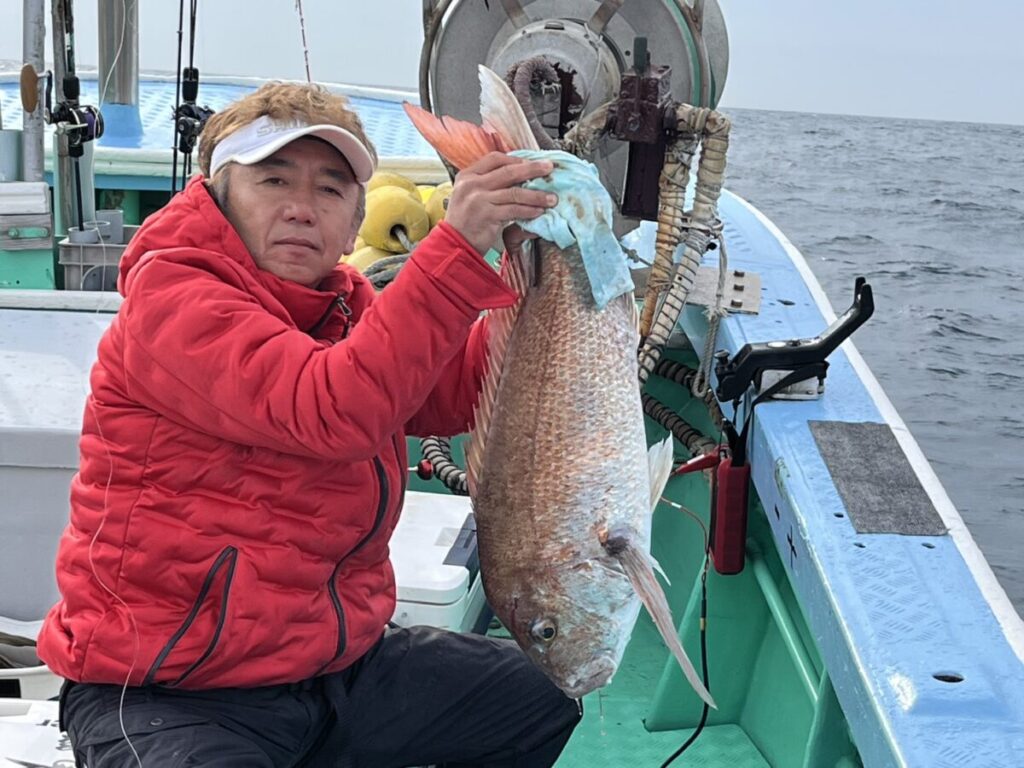 赤いジャケットを着た人物がボートに乗り、片手で大きな魚を持ちながらカメラを見つめている。背景には海と漁具が見える。.