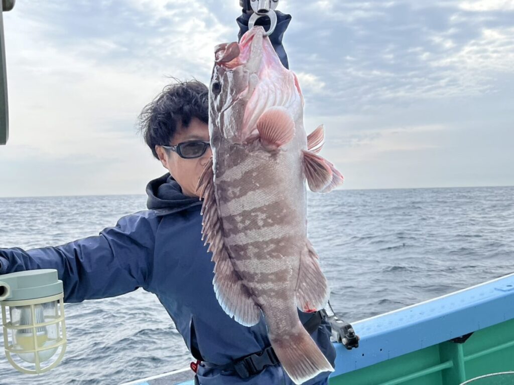 サングラスをかけ、青いジャケットを着た人物が、海と曇り空を背景に、ボートの上で大きな縞模様の魚を掲げている。.