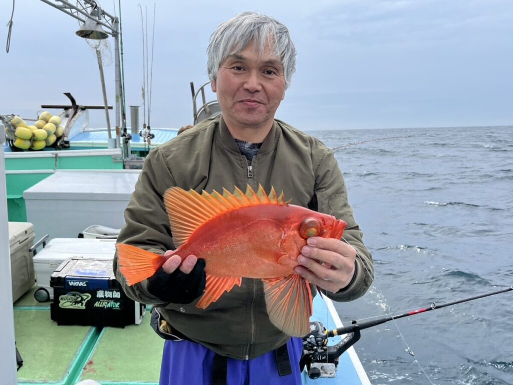 曇り空の下、漁具と海を背景に、ボートの上で鮮やかなオレンジ色の大きな魚を手にした白髪の人物が微笑んでいる。.