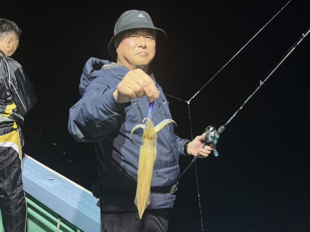 黒っぽい帽子に上着姿の人物が、片手に釣り竿、もう片方の手には釣りたてのイカを持ち、夜の船の上に立っている。.