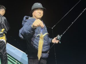 黒っぽい帽子に上着姿の人物が、片手に釣り竿、もう片方の手には釣りたてのイカを持ち、夜の船の上に立っている。.