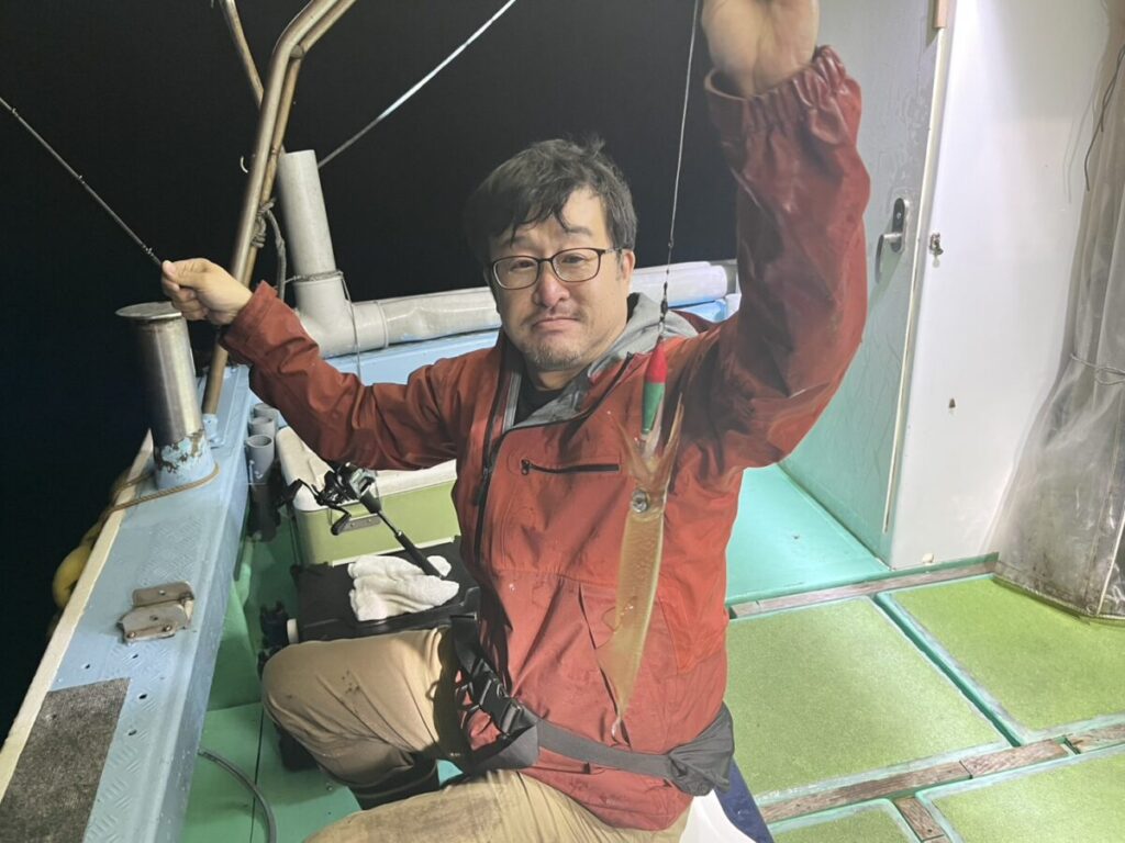 眼鏡をかけ、赤いジャケットを着た男が夜のボートの上で釣り糸を垂れている。彼はカメラ目線で、人工照明の下で釣りをしているように見える。.