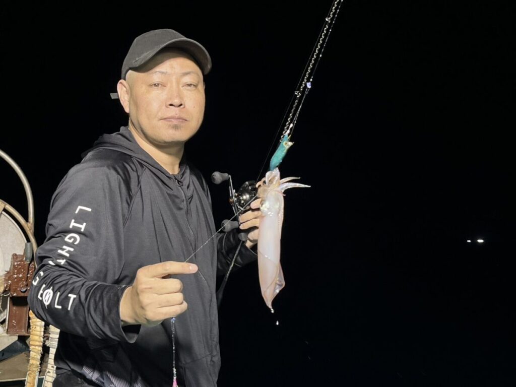 黒い帽子にジャケット姿の男が釣り竿を持ち、釣りたての夜イカを見せる。背景は暗く、男はカメラの方を向いている。.
