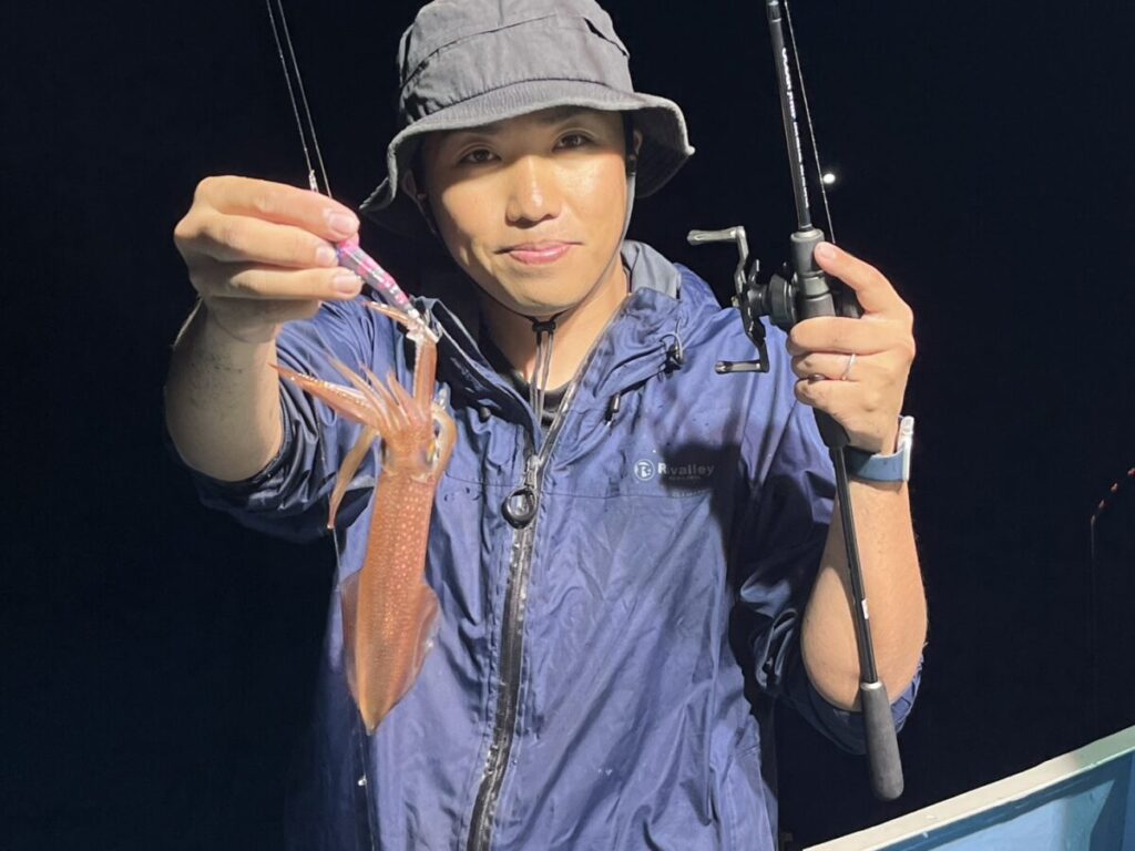 グレーのバケットハットをかぶり、ブルーのジャケットを着た人が、片手に釣り竿を持ち、もう片方の手でエギにかかった小さなイカをアピールしている。.