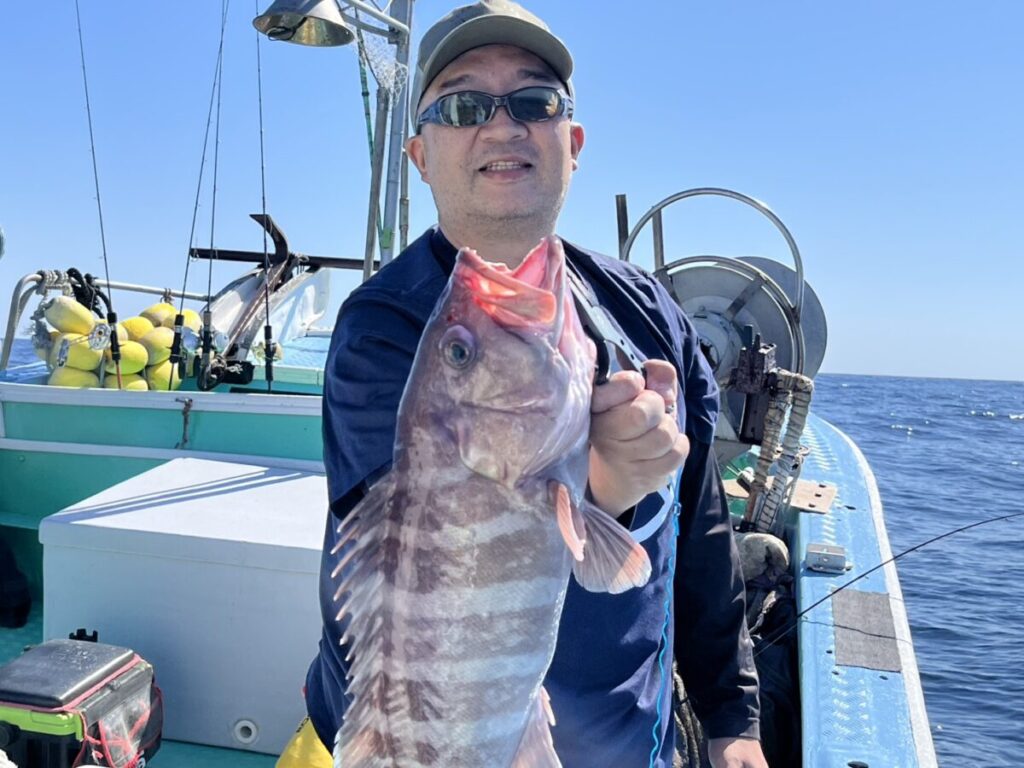 サングラスをかけ、帽子をかぶった人が、青い漁船の上で大きな縞模様の魚を掲げている。.