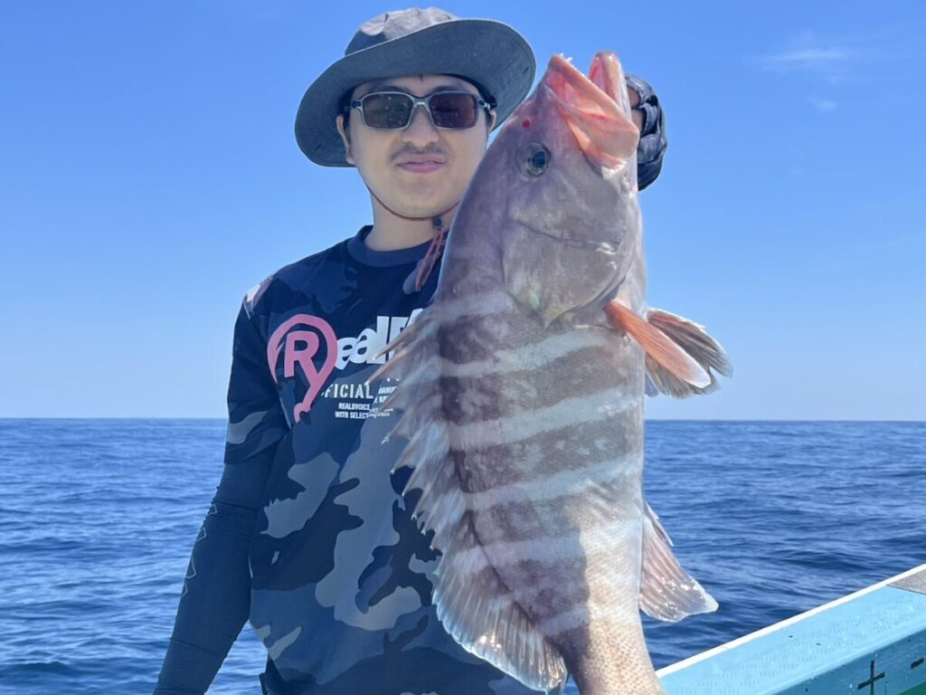 海と青空を背景に、サングラスと帽子をかぶった人物が船の上で大きな縞模様の魚を掲げている。.