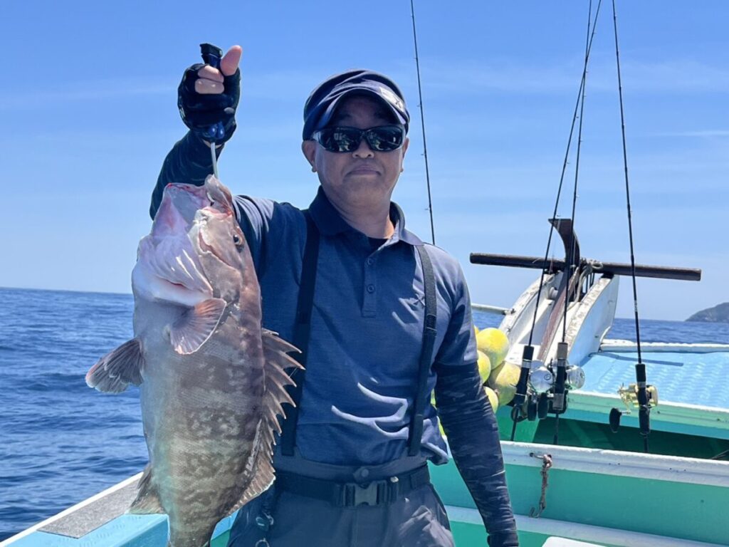 サングラスをかけ、帽子をかぶった人物がボートの上で大きな魚を掲げている。.