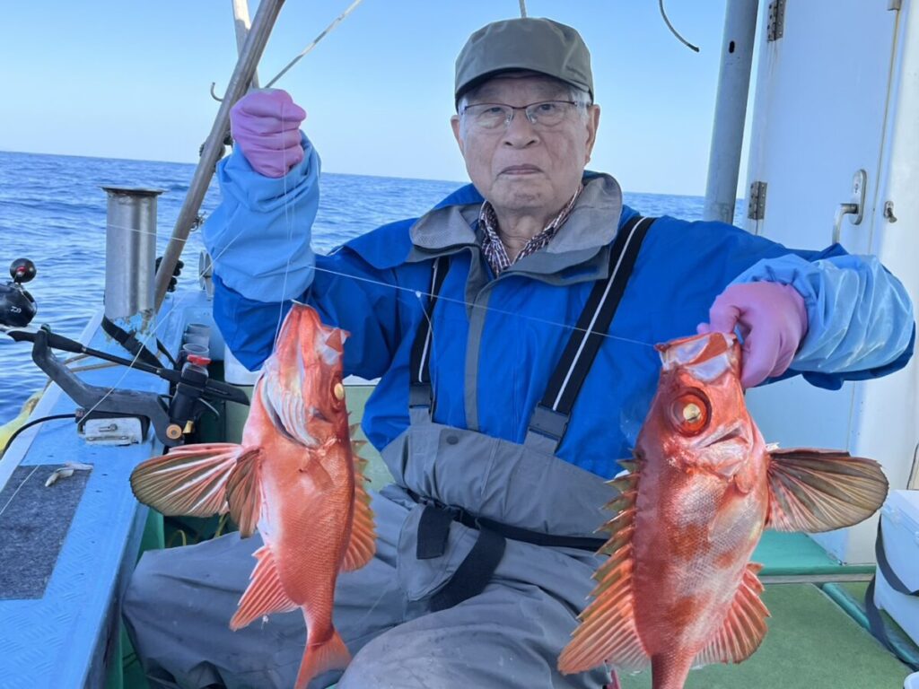 ボートに乗った老人が、鮮やかなオレンジ色の大きな魚を2匹、両手で掲げている。青いジャケットにグレーのオーバーオール、カーキ色の帽子をかぶり、ピンクの手袋をはめている。背景には海と空が見える。.