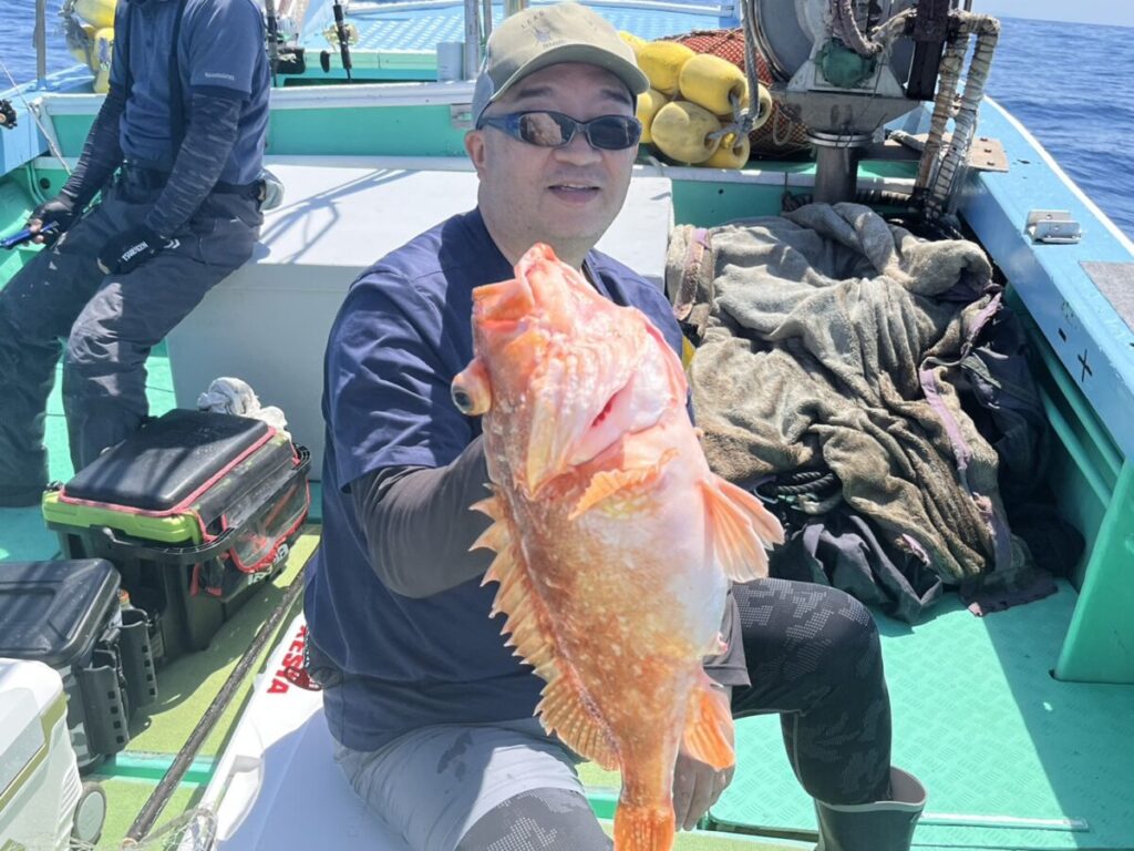 ボートに乗った男がオレンジ色の大きな魚をカメラに向かって掲げ、微笑んでいる。背景には漁具や網が見え、遠くには海と青空が広がっている。.