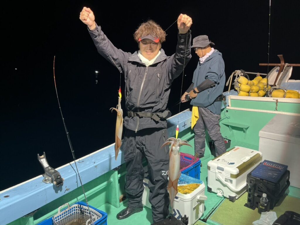 夜のボートに2人が乗っている。手前の一人は微笑みながら2本の釣り竿を掲げ、それぞれにイカを釣り上げている。後ろでもう一人が釣り道具を準備している。.