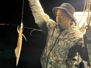 迷彩柄のジャケットと帽子を身にまとい、イカがかかった釣り竿を持つ人物。.