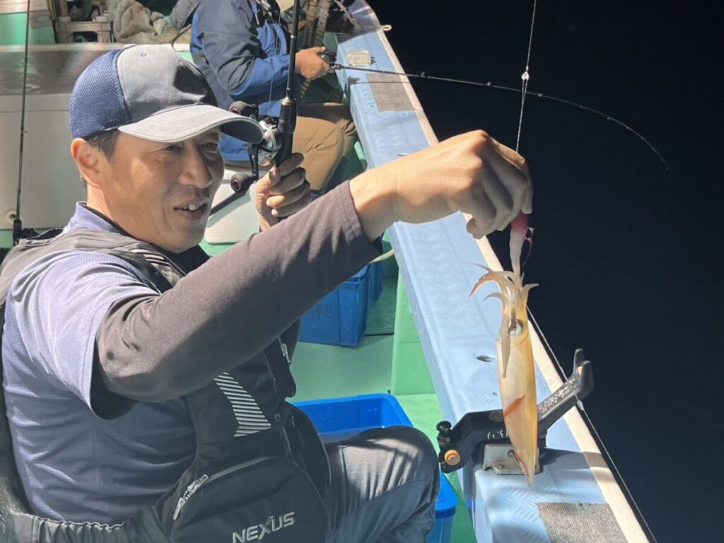 帽子をかぶり、救命胴衣を着た男性が、夜のボートで釣りをしているときに釣れたばかりのイカを掲げている。背景には漁具ともう一人の人物が見える。.