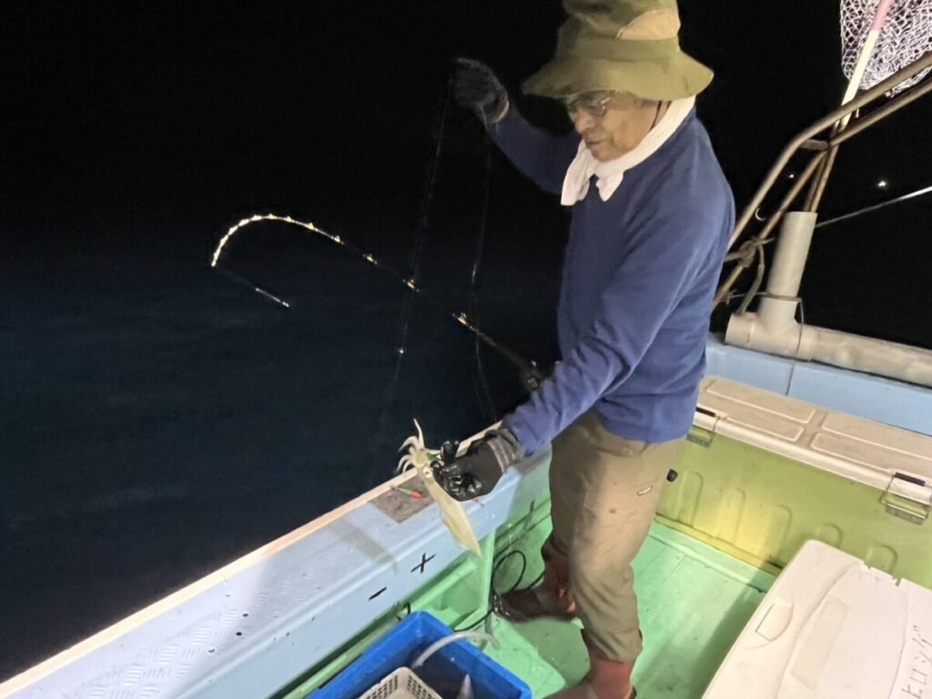 帽子をかぶり、手袋をはめた人が、釣ったばかりのイカを載せた釣り竿を持って夜のボートに立っている。船の甲板には明かりが灯り、漁具が近くに見える。.