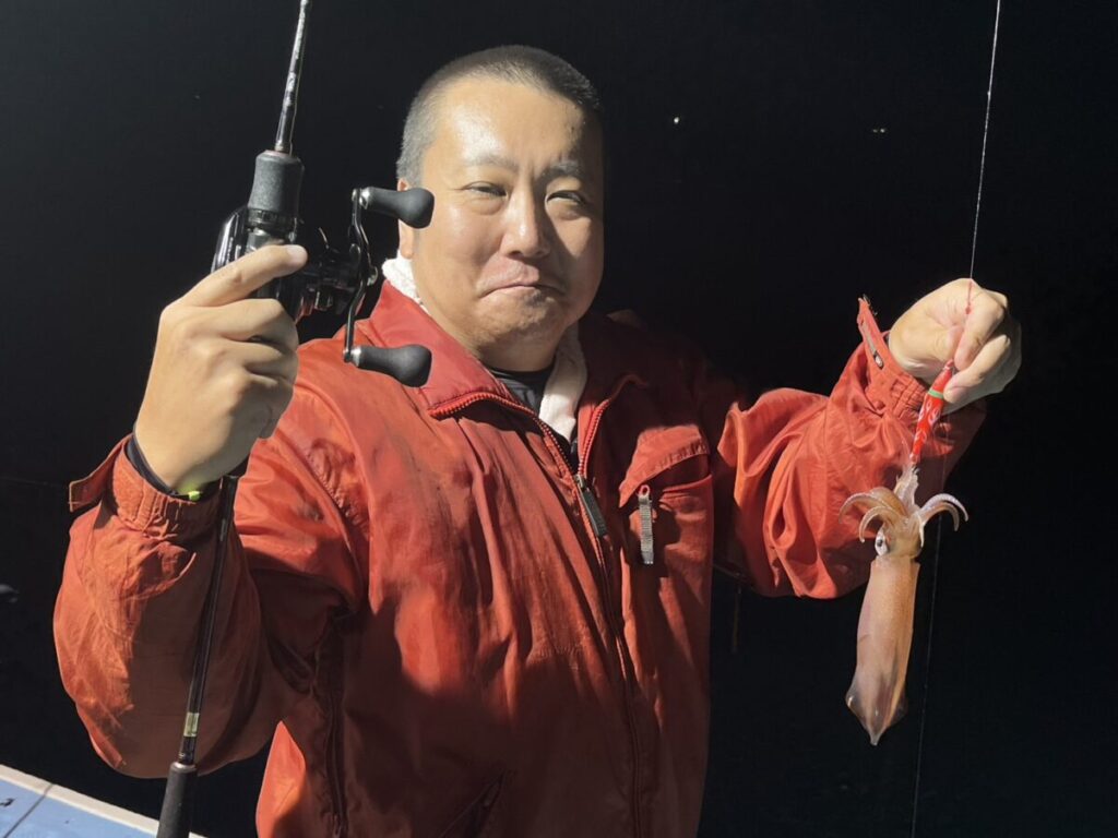 赤いジャケットを着た人が、片手に釣り竿、もう片方の手に小さなイカを持ち、夜の屋外で微笑んでいる。.