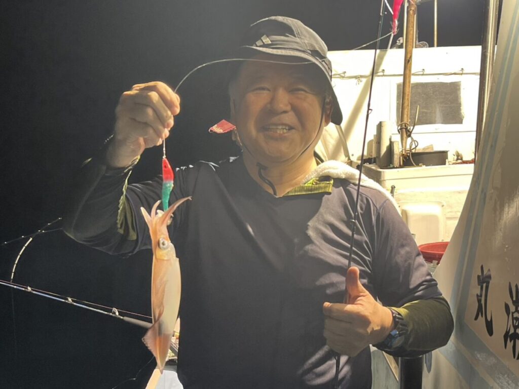 夜の船上で、帽子をかぶった人が釣り糸を垂れたイカを笑顔で掲げている。.