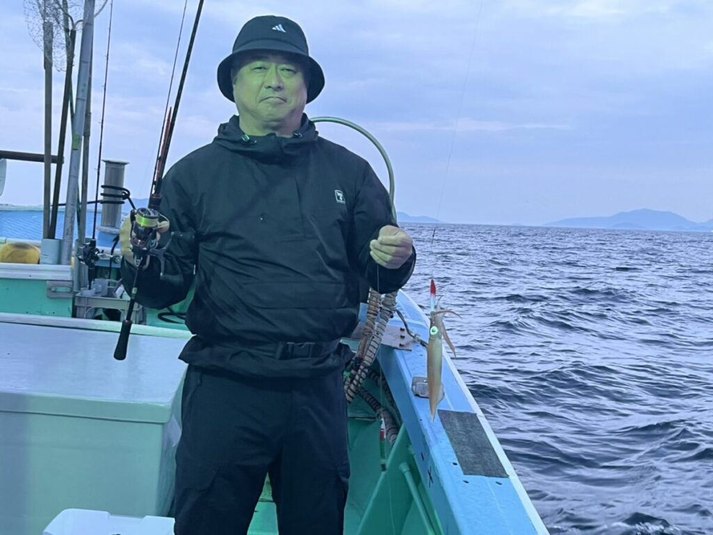 曇り空の下、海と遠くの島々を背景に、黒いジャケットと帽子を被った人物が釣り竿を持ったボートの上に立っている。.