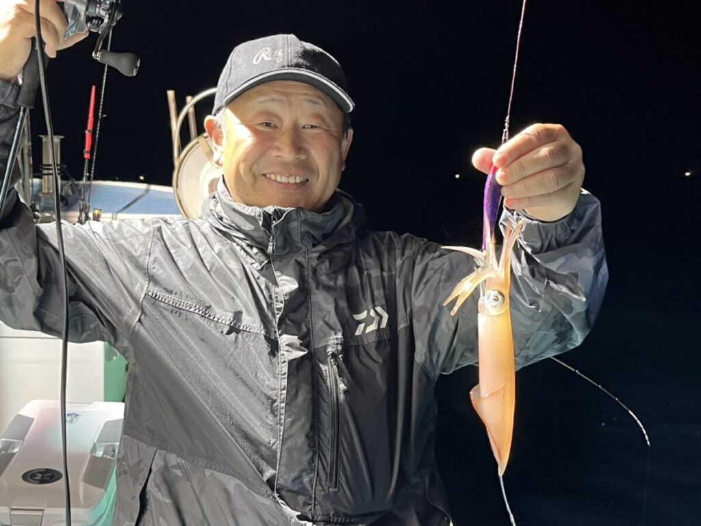 レインジャケットを羽織り、釣り糸を垂れた釣りたてのイカを掲げる笑顔の男。.