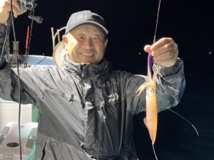 レインジャケットを羽織り、釣り糸を垂れた釣りたてのイカを掲げる笑顔の男。.