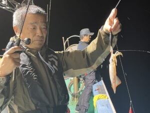 ヘッドセットとライフジャケットを着用した男性が、夜釣り竿に釣りたてのイカを掲げている。.