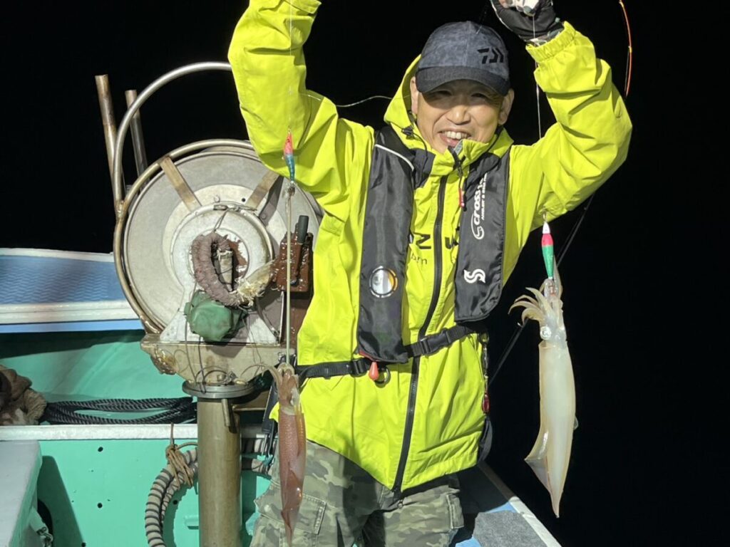 鮮やかな黄色のジャケットを着た笑顔の漁師が、夜、船から獲れたてのイカを誇らしげに2つ掲げている。.