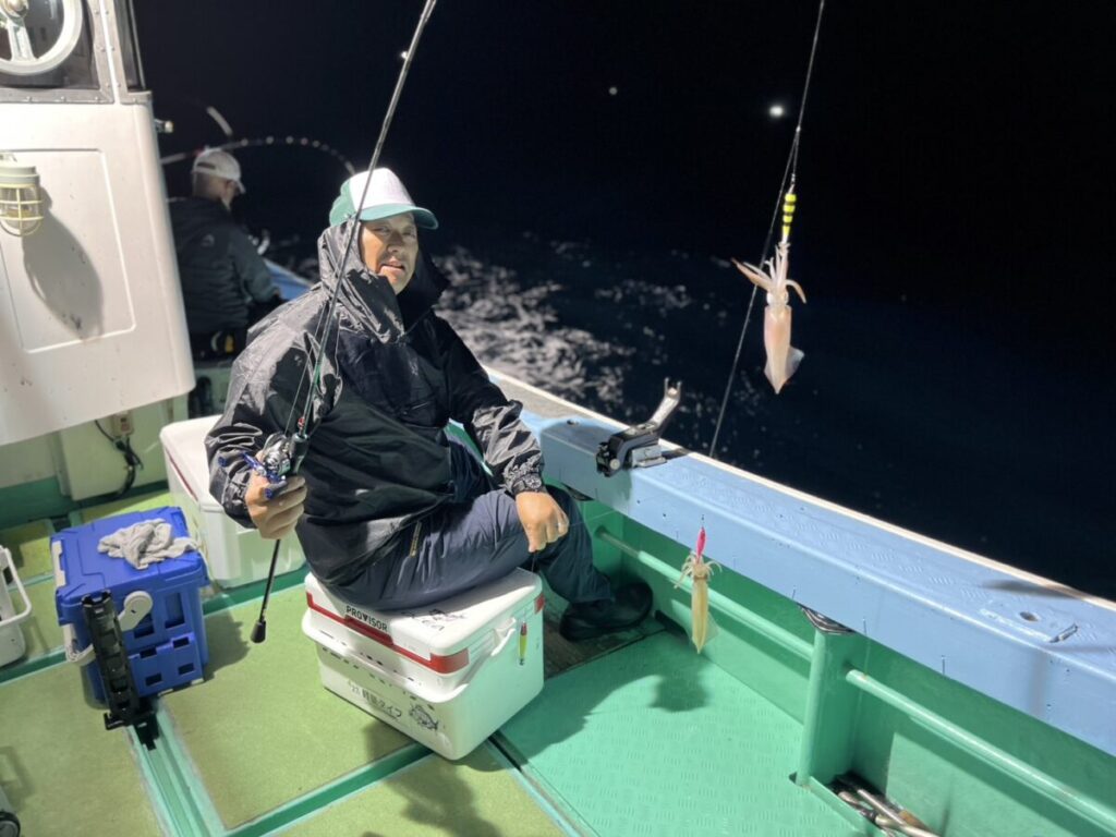 夜の船上で、クーラーの上に座って釣り竿を持つ人。釣り糸には釣れたイカがぶら下がっている。.
