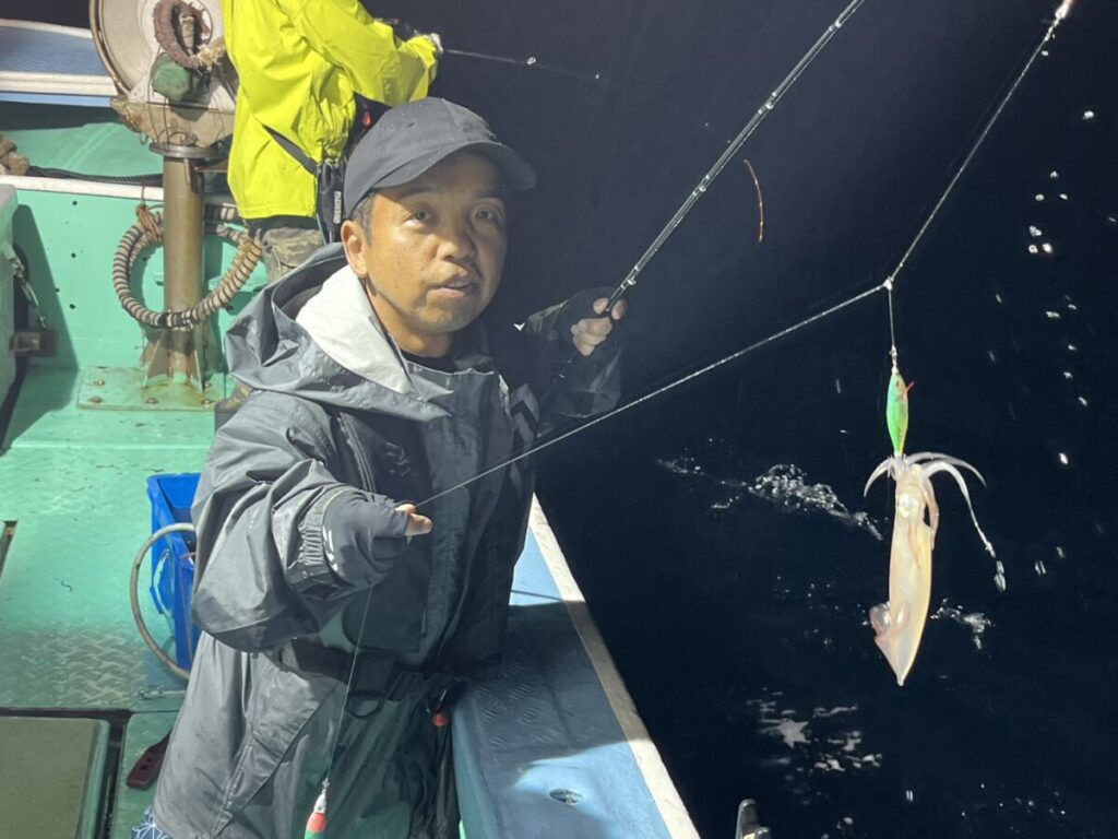 釣り用の上着と帽子をかぶった男が釣り竿を持ち、暗い水面を背景に釣れたイカを緑色のルアーで巻き上げる。.