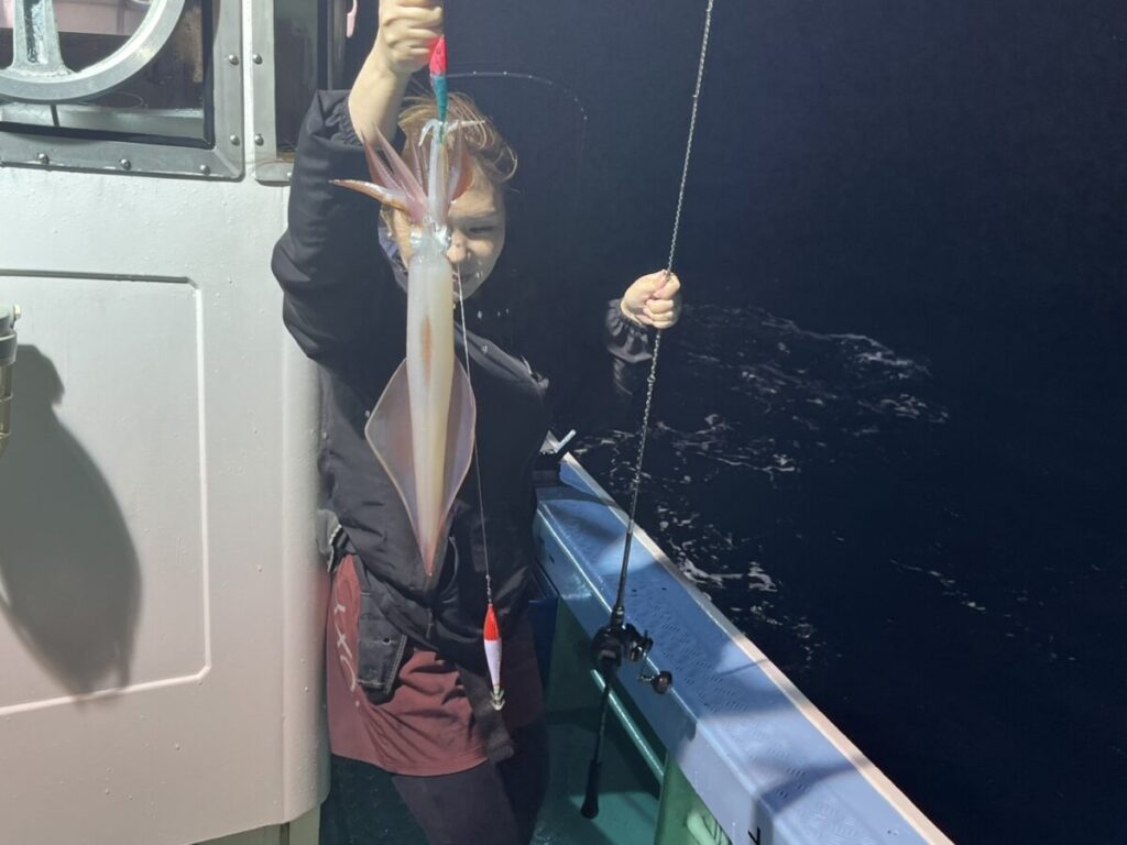 夜の船上で、釣り上げた大きなイカを掲げて誇らしげに笑う人。背景には海と釣り竿が見える。.