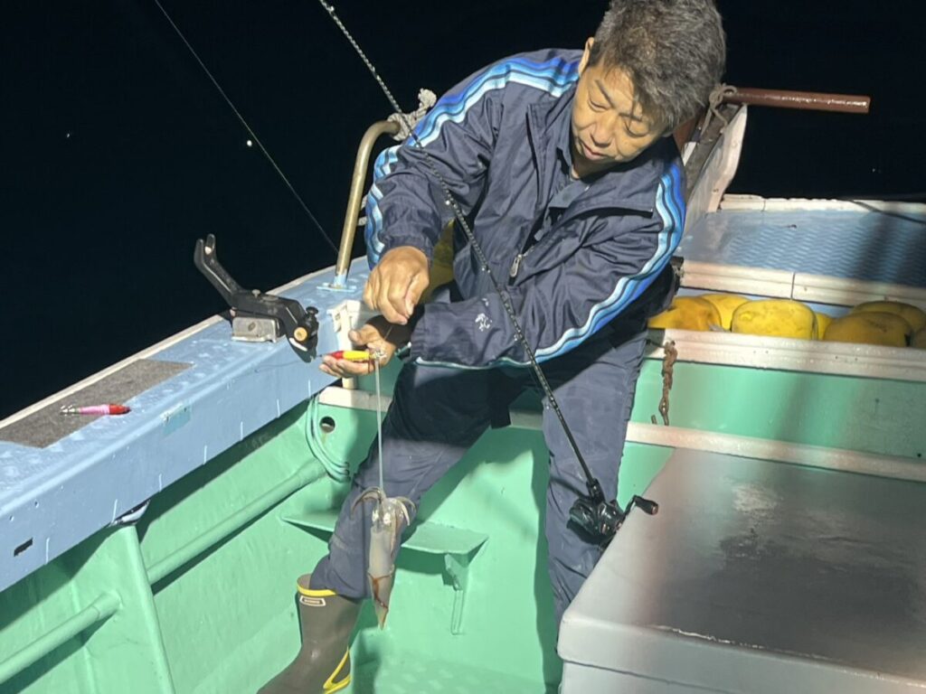青いジャケットに長靴を履いた人物が夜のボートの上に立ち、釣り竿でイカを釣っている。背景には黄色い浮きが見える。.
