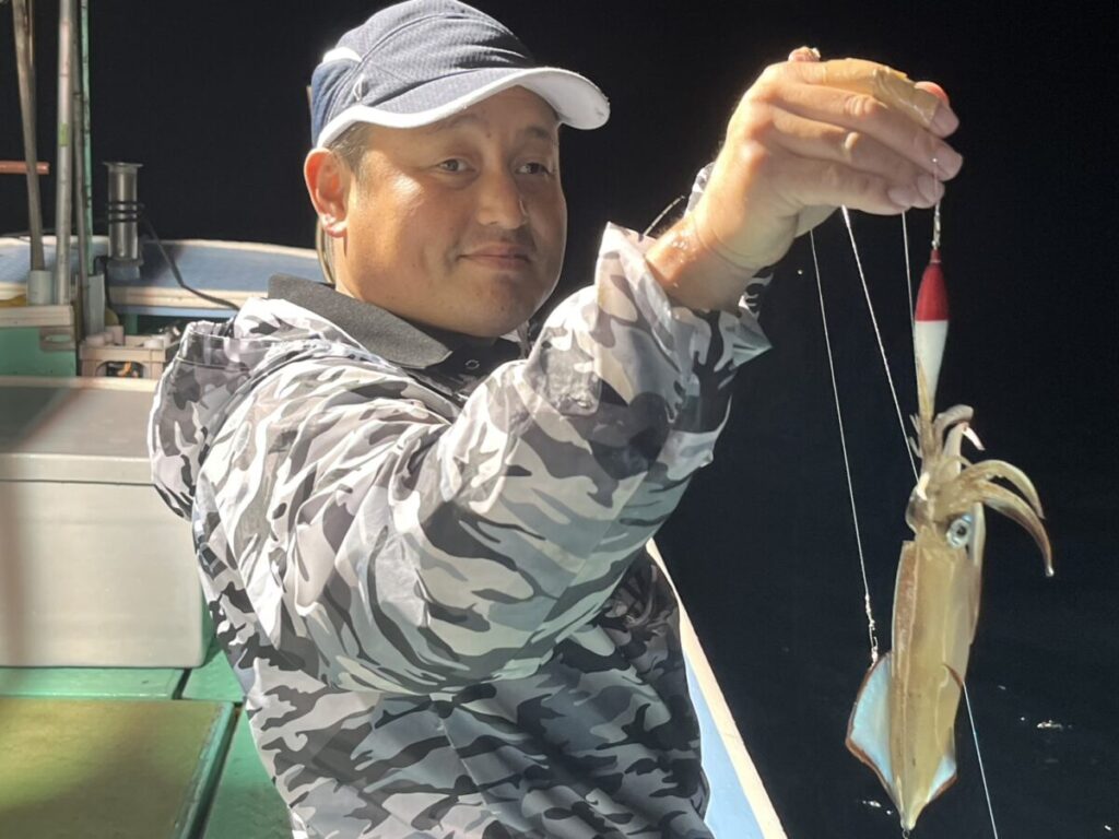 迷彩柄の上着と帽子を身にまとい、夜の船上で釣りたてのイカをエギで釣り上げる。.
